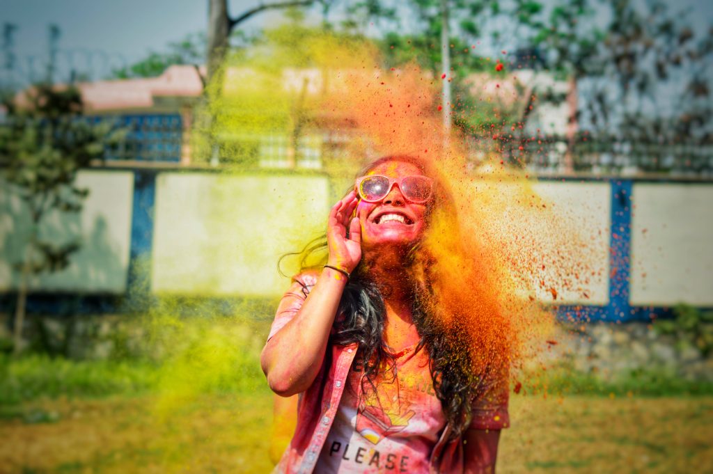 Smiling woman in sunglasses celebrates vibrant Holi festival with bright yellow and orange powder burst Wallpaper