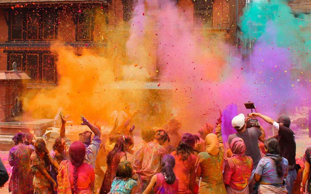 Joyful Holi festival crowd throwing vibrant orange pink teal powder by brick wall wallpaper