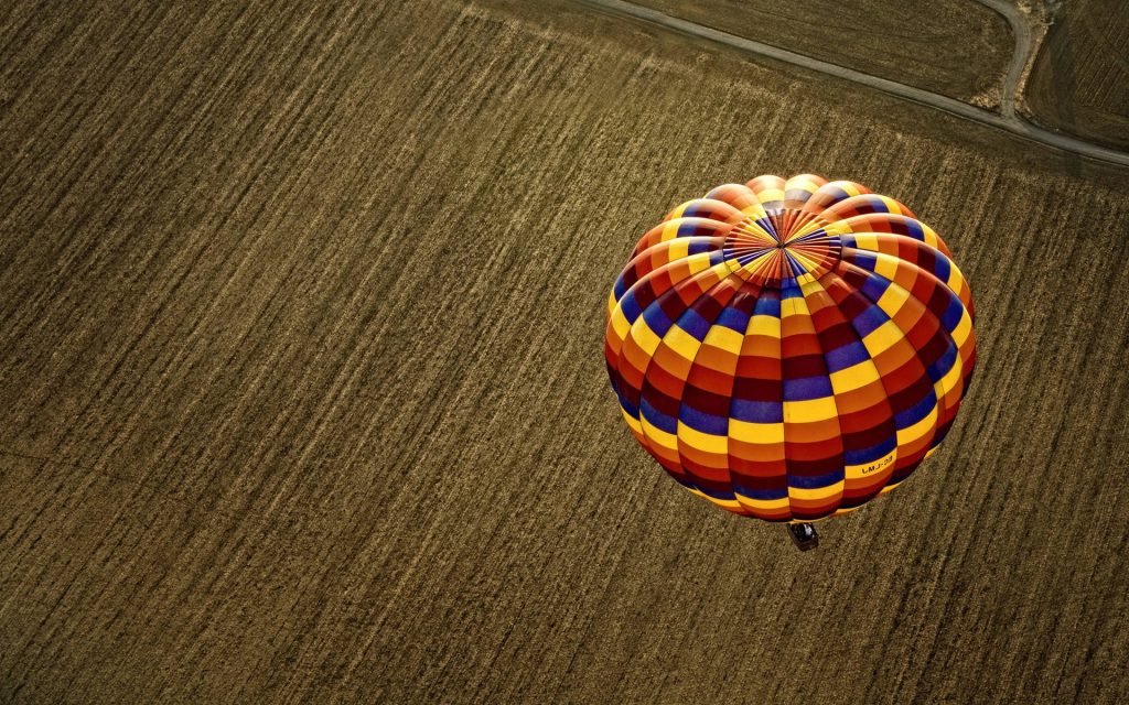 Aerial view of colorful hot air balloon EC-FW soaring over textured brown field wallpaper background
