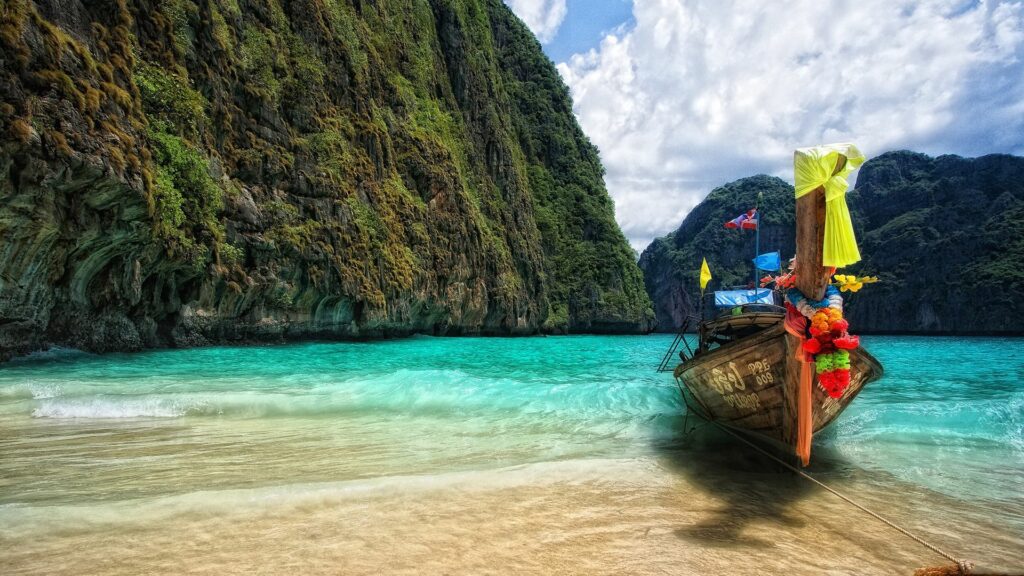 Colorful long-tail boat moored in clear turquoise water by tropical cliffs under cloudy sky Wallpaper
