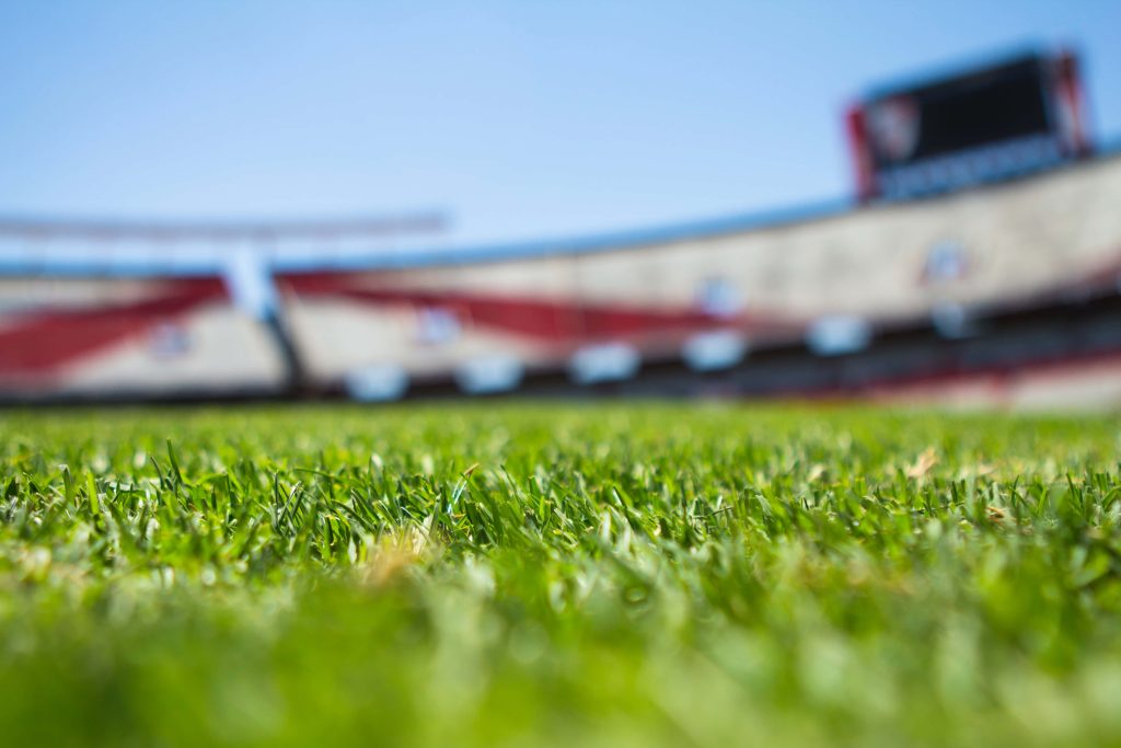 Low-angle vibrant green turf football stadium with blurred red white bleachers and clear sky Wallpaper