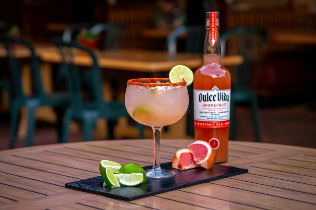 Vibrant Margarita with lime chili rim and Dulce Vida Grapefruit bottle on wooden table decor Wallpaper