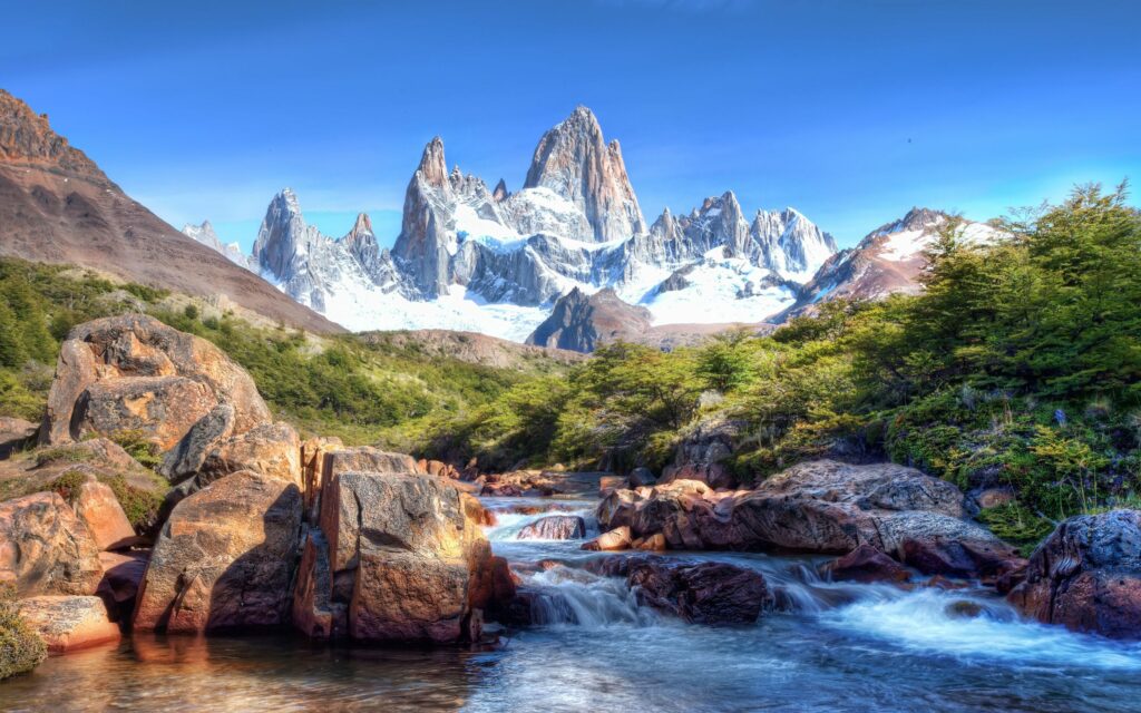 Clear river flowing over rocks with green forest and snow-capped mountains under blue sky wallpaper