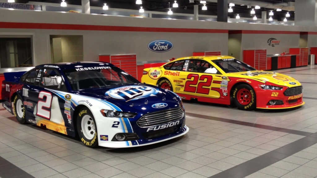 Two colorful NASCAR Ford race cars parked side by side showing sponsor logos in modern garage Wallpaper