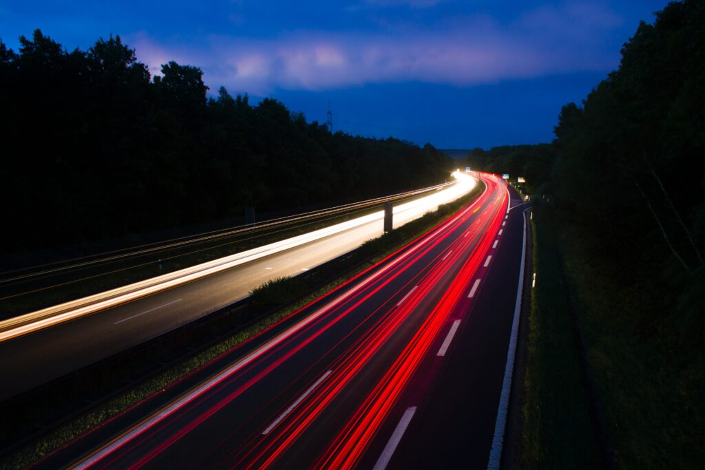 Curving road at night with red and white light trails under deep blue sky and dark trees wallpaper