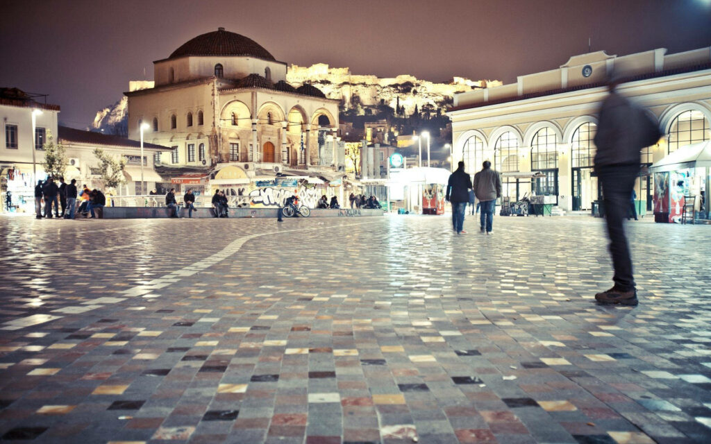 Nighttime plaza in Athens with historic buildings and illuminated Acropolis bustling with people Wallpaper