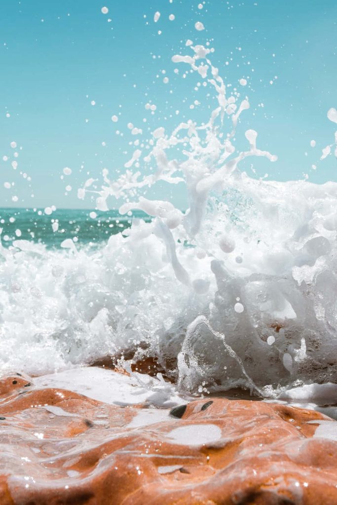 Vibrant ocean wave crashing on reddish coastal rocks with white foam and turquoise sea background Wallpaper
