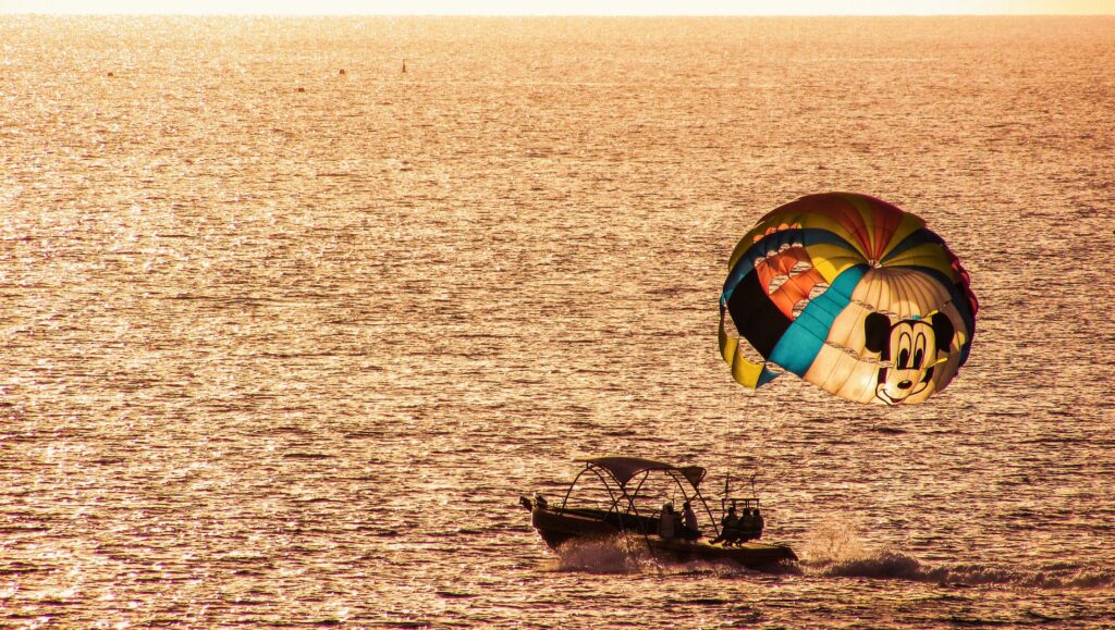 Parasailing over shimmering sea at sunset with colorful cartoon parachute and boat adventure wallpaper