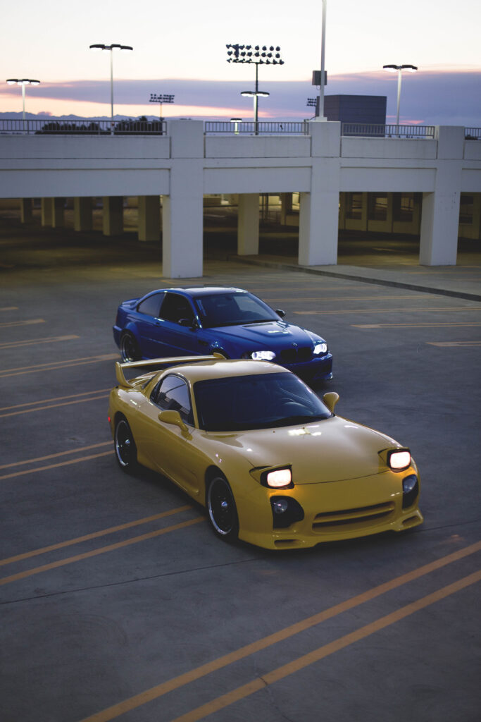 Vibrant Parking Structure: Yellow Mazda RX-7 & Blue BMW at Sunset Wallpaper