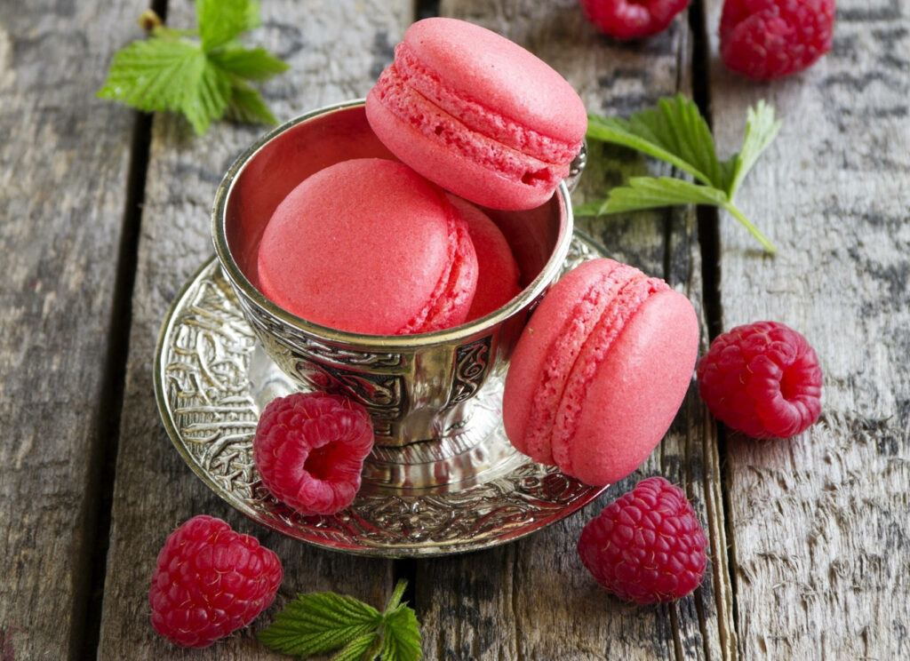 Vibrant pink macarons and fresh raspberries in ornate silver cup on rustic wooden background wallpaper