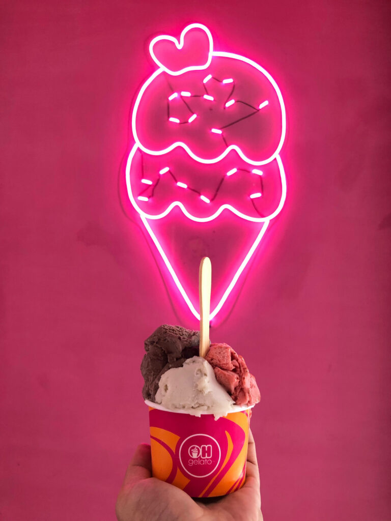 Vibrant pink wallpaper with neon ice cream cone sign and colorful gelato cup in hand