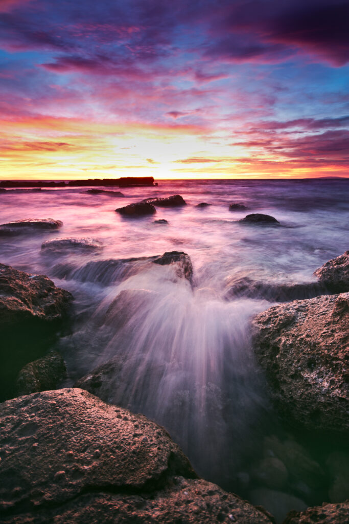 Sunset sky in pink purple and orange hues over sea with waves crashing on rugged coastal stones Wallpaper