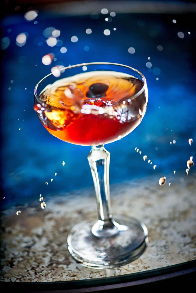 Pisco Manhattan cocktail in coupe glass with dark cherry garnish and vibrant amber hues lighting Wallpaper