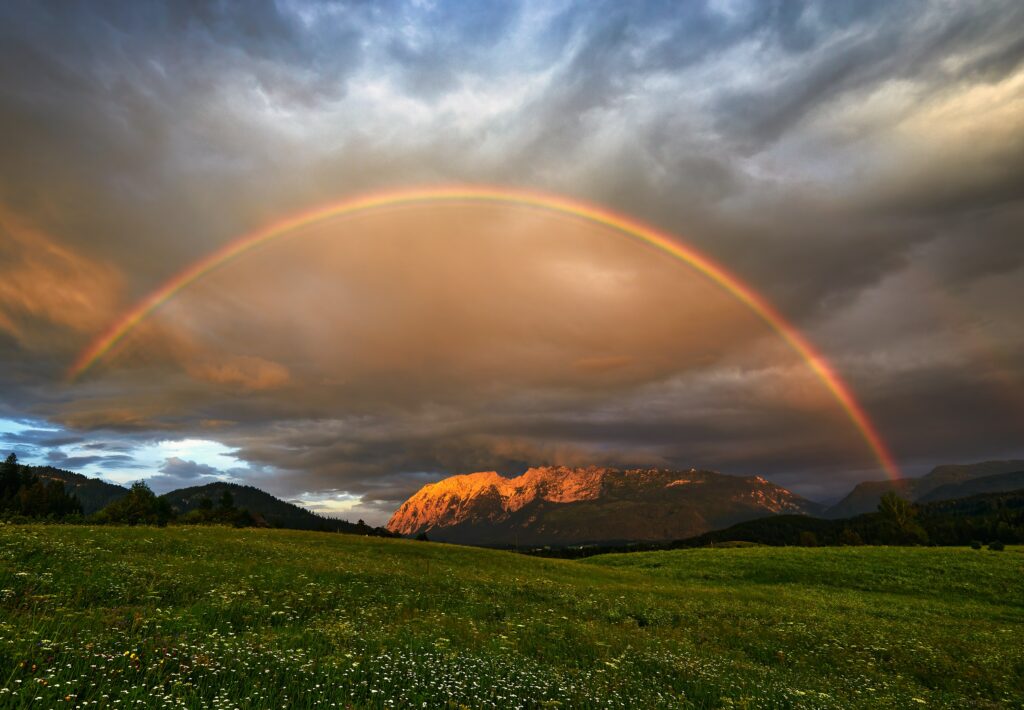 Vibrant rainbow over green meadow with wildflowers and sunlit mountains dramatic sky wallpaper
