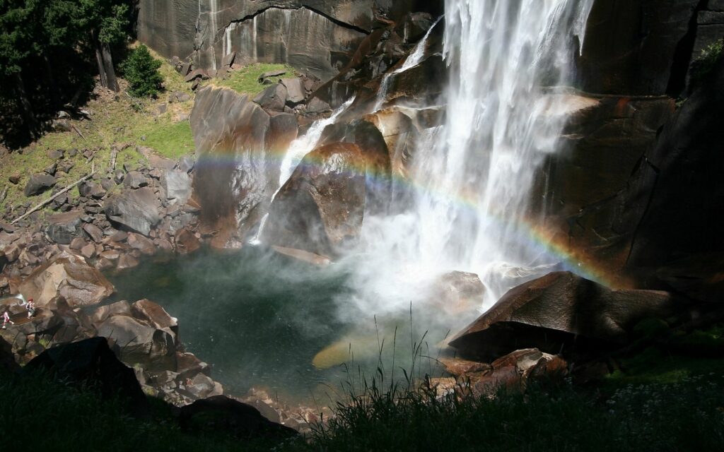 Powerful waterfall over rocky stones with rainbow mist and sunlight highlighting lush greenery Wallpaper