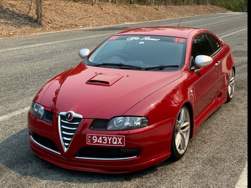 Red Alfa Romeo GT parked on open road with scenic nature and clear blue sky background Wallpaper