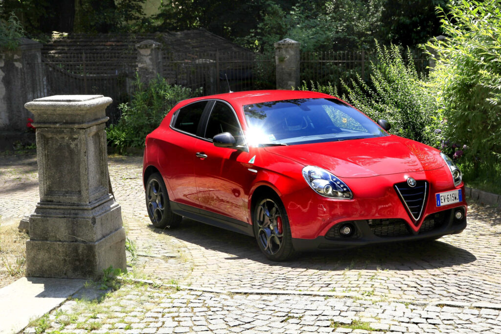Red Alfa Romeo Giulietta parked on sunlit cobblestone path with lush green background wallpaper