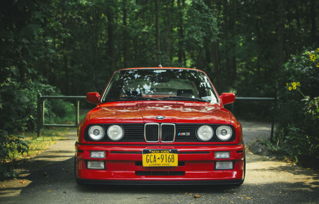 Red BMW E30 M3 classic front view parked on shaded forest driveway automotive wallpaper background