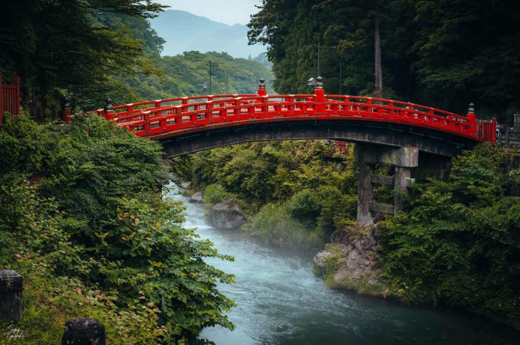 Vibrant red bridge over flowing river with mist and lush green trees in serene natural landscape Wallpaper