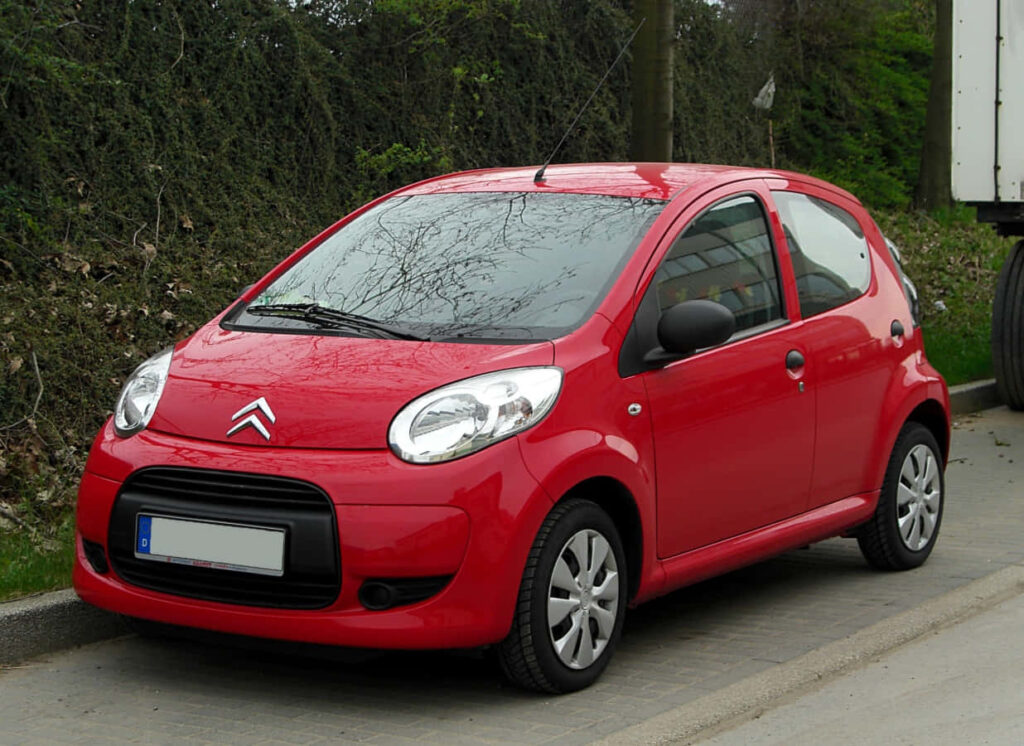 Vibrant Red Compact Car Wallpaper Surrounded by Lush Greenery