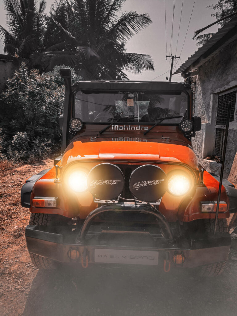 Red Mahindra Thar with glowing headlights parked outdoors by trees and rustic building in 4K Wallpaper