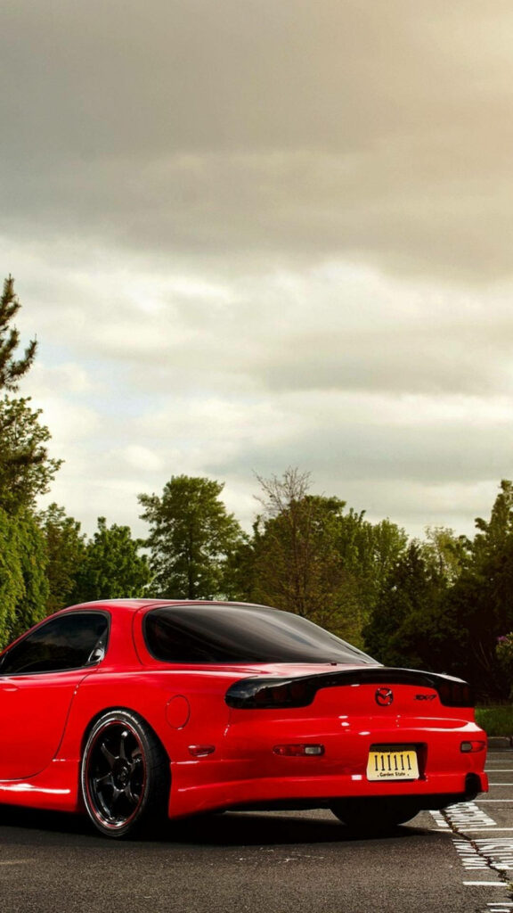 Red Mazda RX-7 sports car parked on quiet street with green trees and cloudy sky background Wallpaper