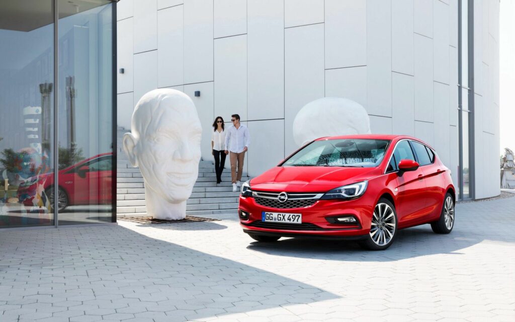 Vibrant Red Opel Astra K in Urban Landscape with Modern Sculptures Wallpaper