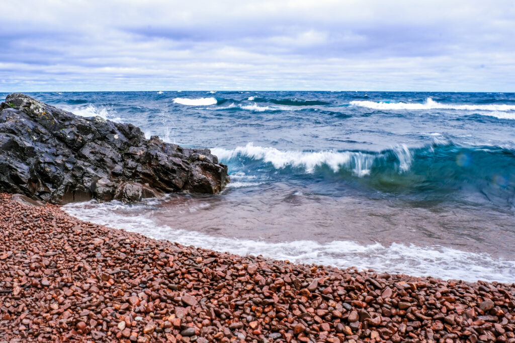 Vibrant seascape with deep blue waves crashing on rugged stones and smooth reddish pebbles Wallpaper
