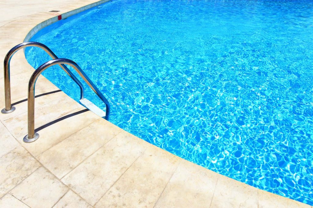 Stainless steel pool ladder with sparkling turquoise water and beige tiled deck on sunny day Wallpaper