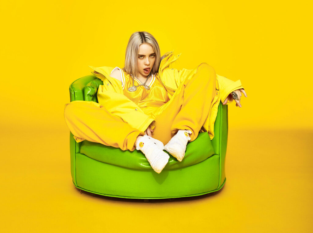 Vibrant Billie Eilish: A Captivating 4k Portrait of the American Singer Rocking Yellow Attire, Relaxing on a Green Sofa with a Brilliant Yellow Backdrop Wallpaper