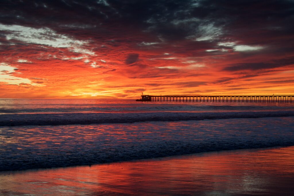Sunset over calm sea with vibrant orange sky and pier extending into peaceful coastal horizon Wallpaper