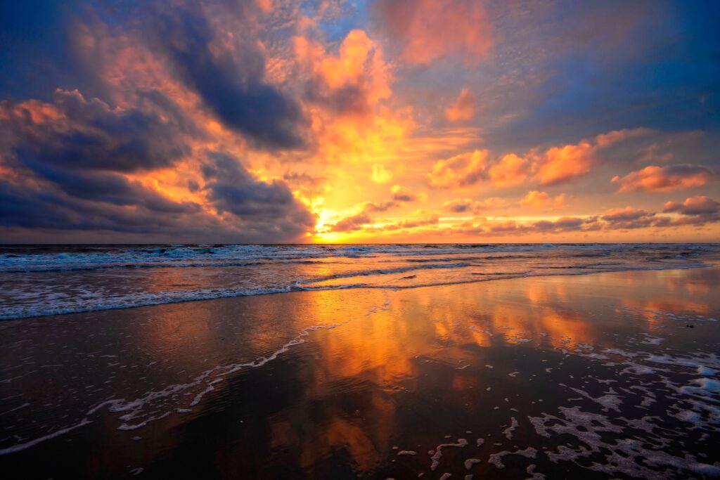 Vibrant orange and purple sunset sky reflecting on wet sand by calm sea with drifting clouds Wallpaper