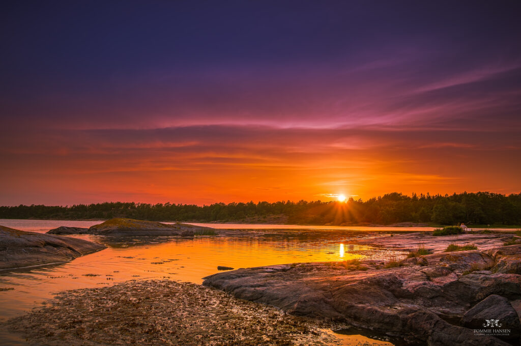 Vibrant sunset over river with golden reflections rocky shore silhouetted trees purple orange sky Wallpaper