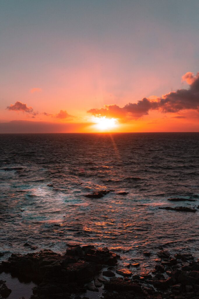 Vibrant sunset over sea with glowing sun orange red hues reflecting on water dark stones foreground Wallpaper