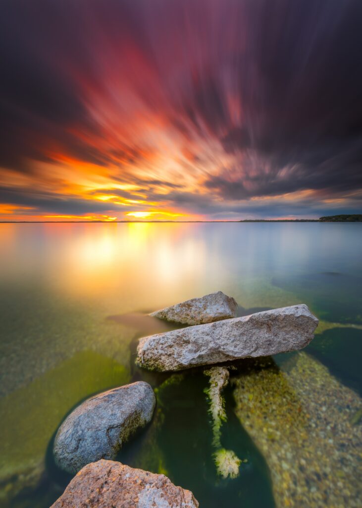 Vibrant sunset over calm sea with textured shore stones in surreal long-exposure wallpaper background