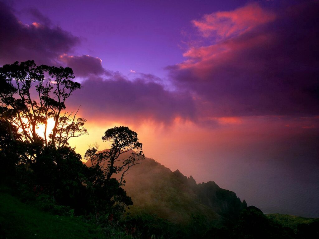 Sunset wallpaper with dramatic purple orange sky silhouetted trees and rugged hillside landscape