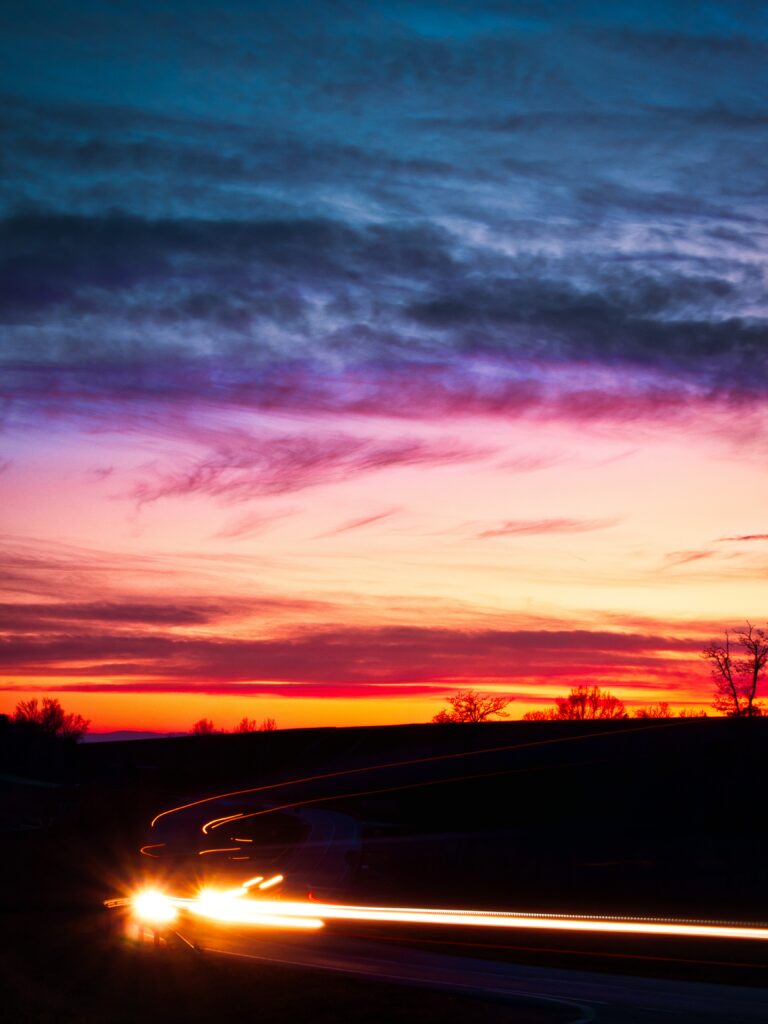 Dramatic sunset sky with purple orange hues and car light trails on winding rural road wallpaper