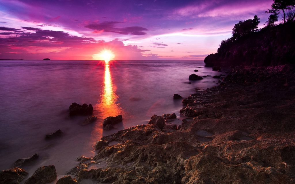 Vibrant sunset with purple magenta sky golden light on ocean and rugged rocky shore wallpaper background
