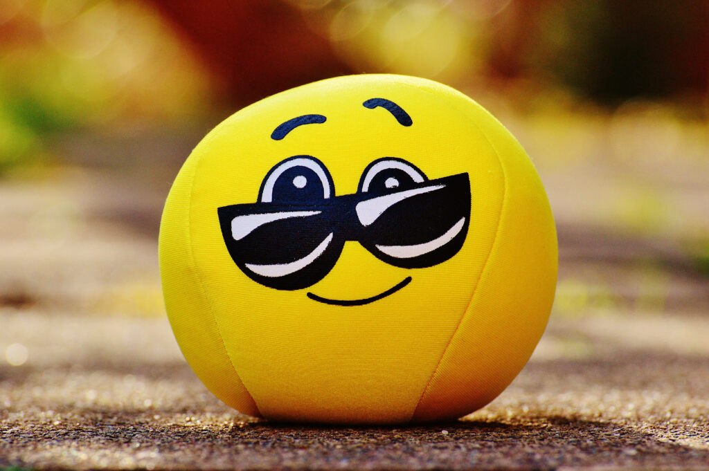 Sunny Smiley: Vibrant Yellow Emoji Plush With Cool Sunglasses on a Colorful Background Wallpaper