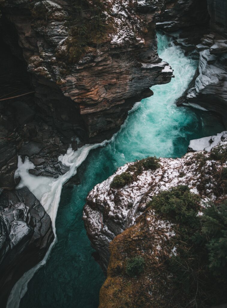 Aerial view of turquoise river flowing through rugged cliffs with greenery and snow patches wallpaper