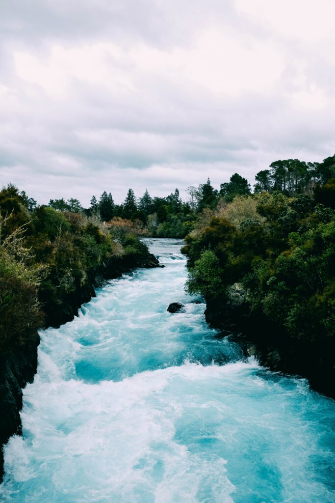 Vibrant turquoise river with foamy waves flowing through dense green forest under cloudy sky wallpaper