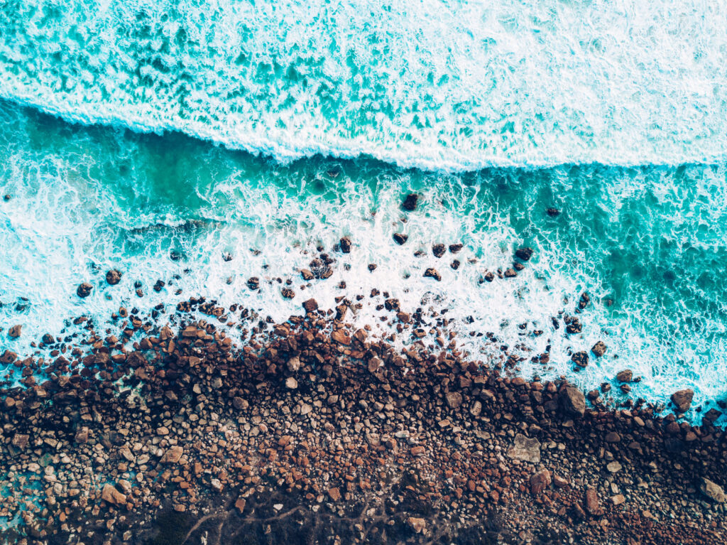Aerial view of turquoise waves crashing on rugged rocky shoreline with foamy surf and pebbles Wallpaper