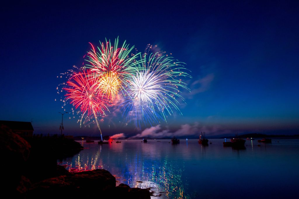 Vibrant USA fireworks over calm harbor with colorful reflections and silhouetted boats at night Wallpaper