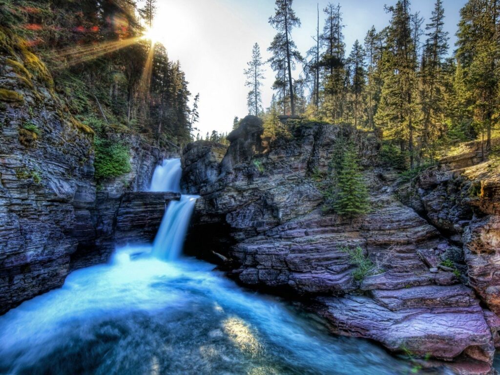 Vibrant waterfall flowing through rocky cliffs with sunlight and tall pine trees landscape background Wallpaper