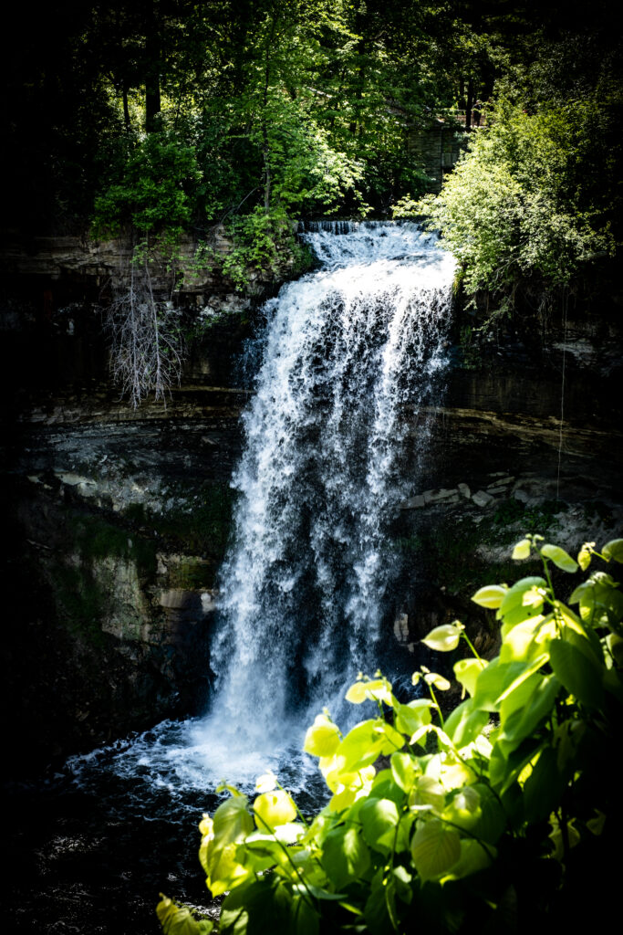 Vibrant waterfall cascading over rocks with lush green plants under sunlight in serene nature background Wallpaper