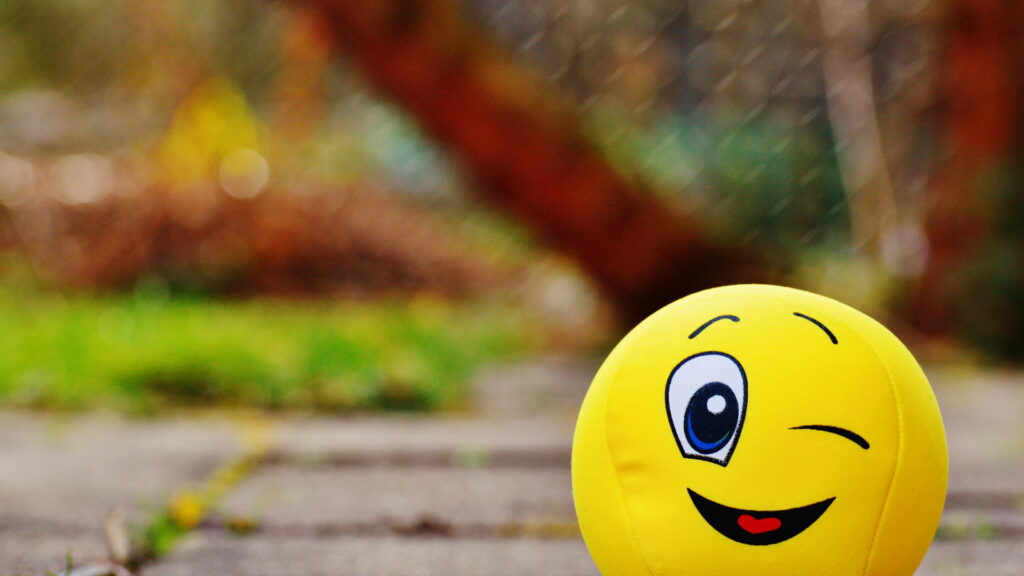 Vibrant HD wallpaper of a cheerful yellow winking emoji plush with soft outdoor bokeh background