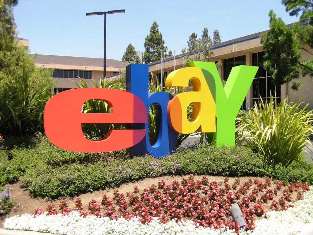 Vibrant eBay logo sculpture outdoors with lush greenery flowers blue sky modern office background Wallpaper