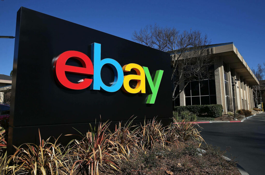 eBay multicolored logo on black sign outside modern glass brick building with blue sky and greenery Wallpaper