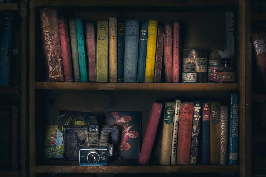 Vintage camera on bookshelf with colorful old novels creating nostalgic artistic wallpaper background
