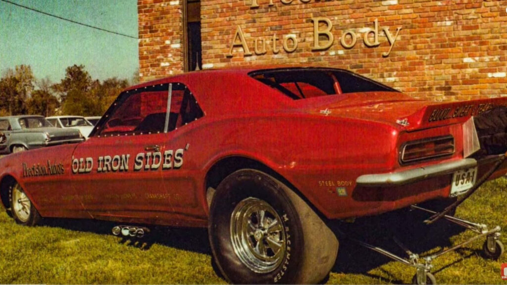 Vintage red Old Iron Sides drag racing car with oversized tires parked outside brick shop Wallpaper